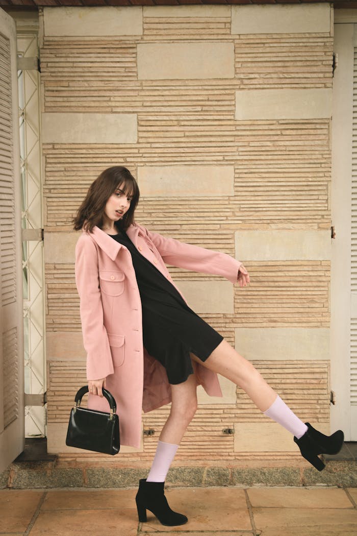 why-choose-us Fashion model in pink coat and black dress striking a dynamic pose indoors with stylish black heels.